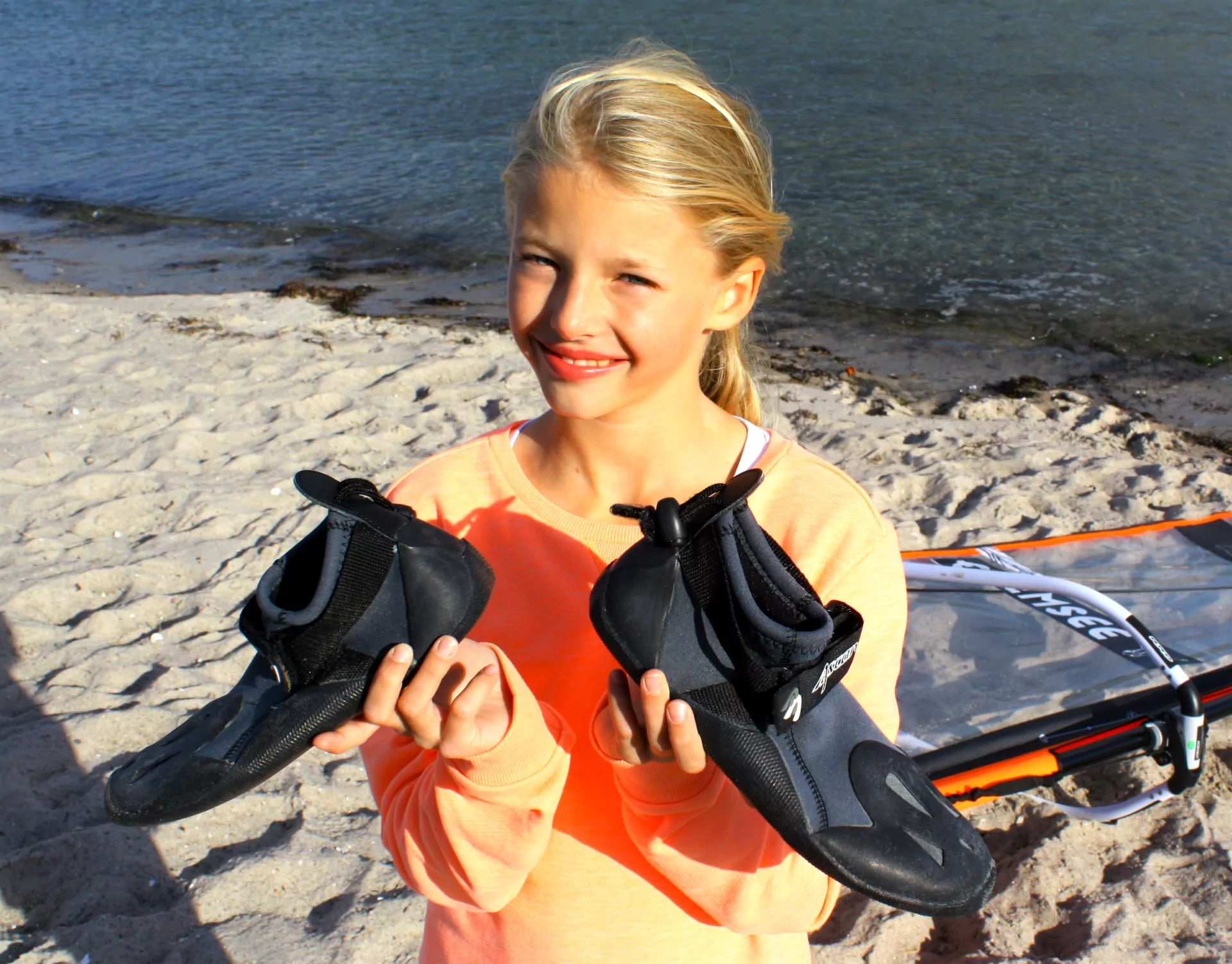 Lächelndes Mädchen am Strand hält Neoprenschuhe, Wassersportausrüstung liegt im Sand