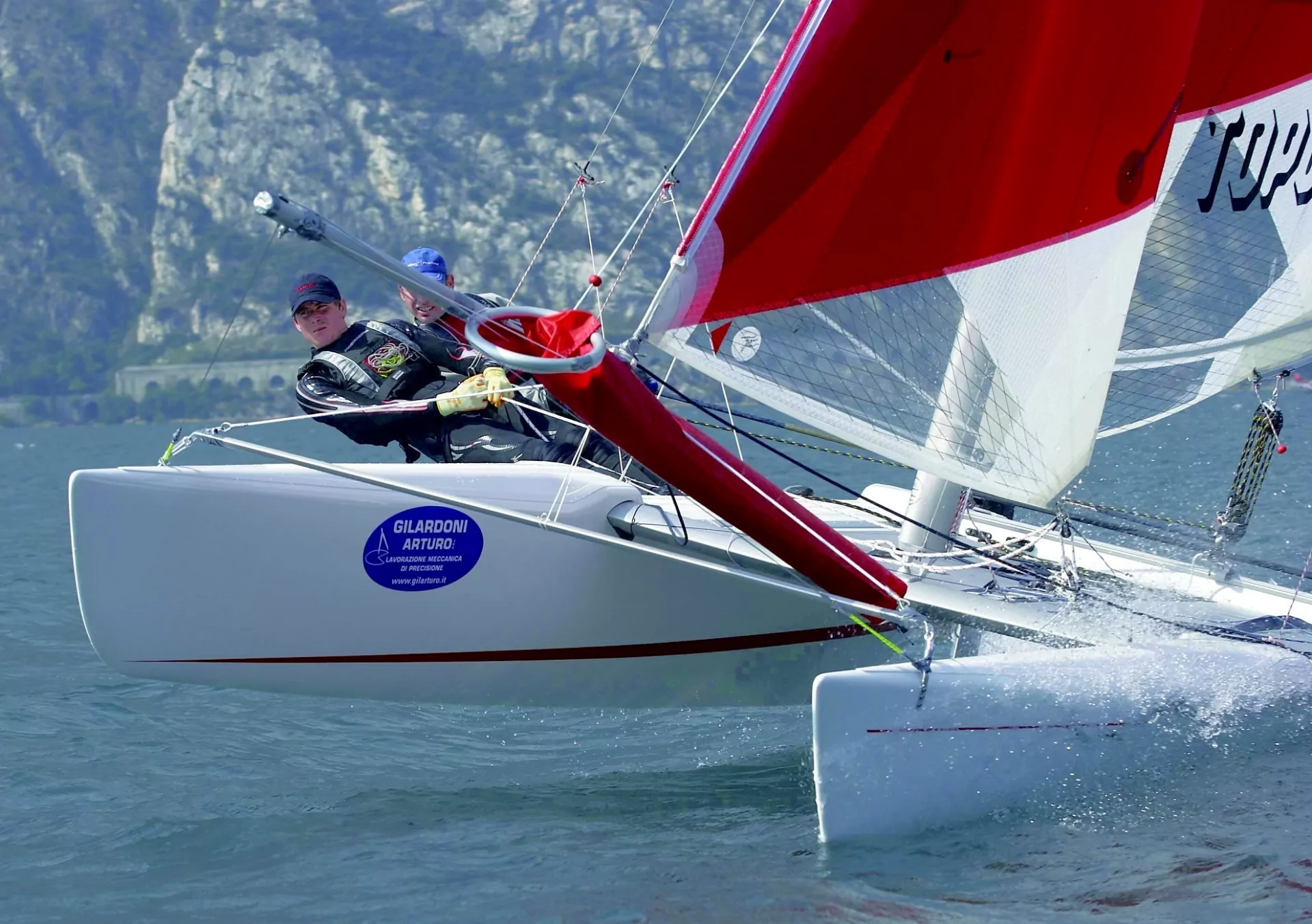 Segelkatamaran mit rotem Segel, zwei Segler lehnen aus, Spritzwasser vor Bergkulisse, dynamisches Regatta-Bild