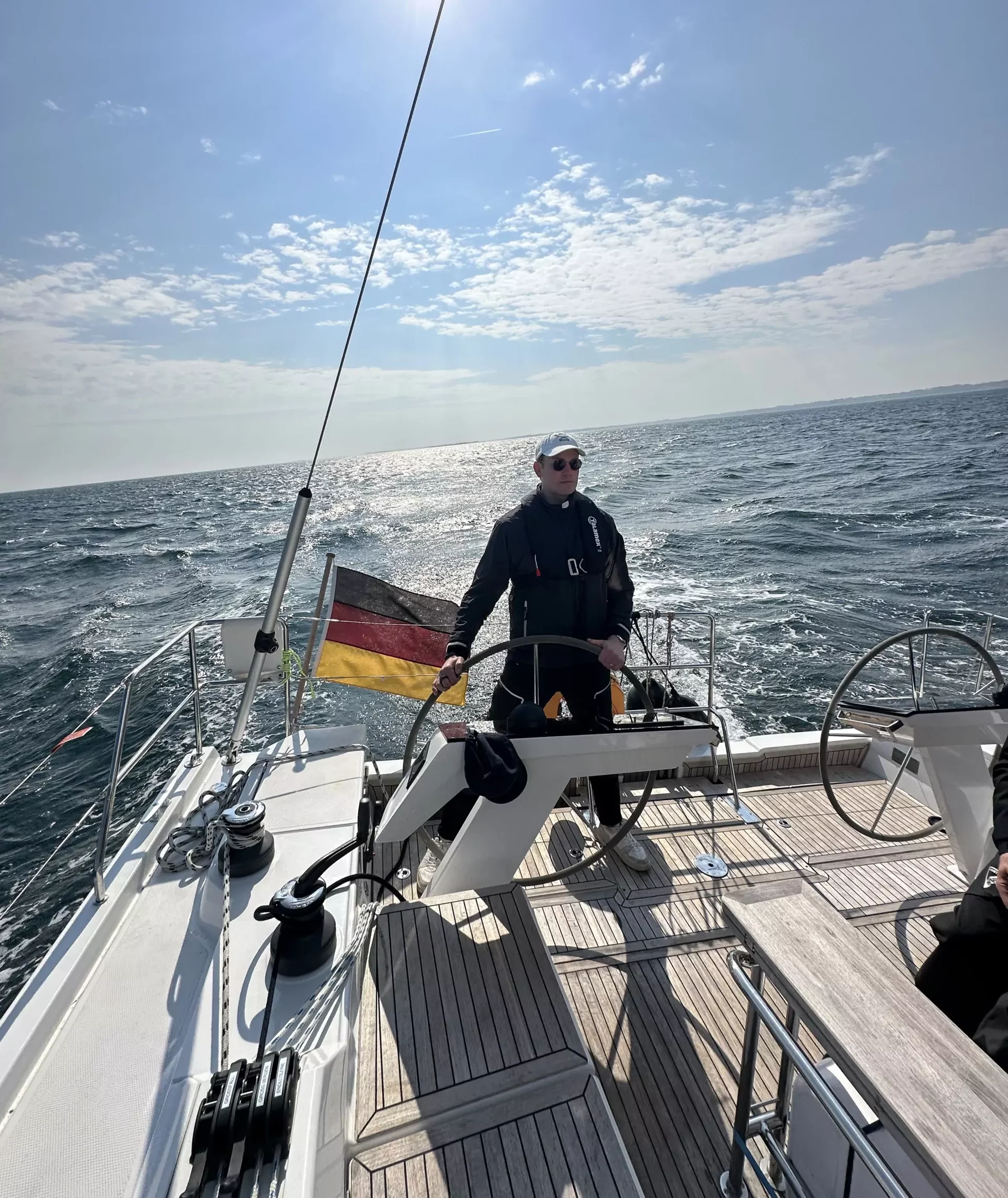 Mann am Steuer einer Segelyacht mit deutscher Flagge, offenes Meer und sonniger Himmel