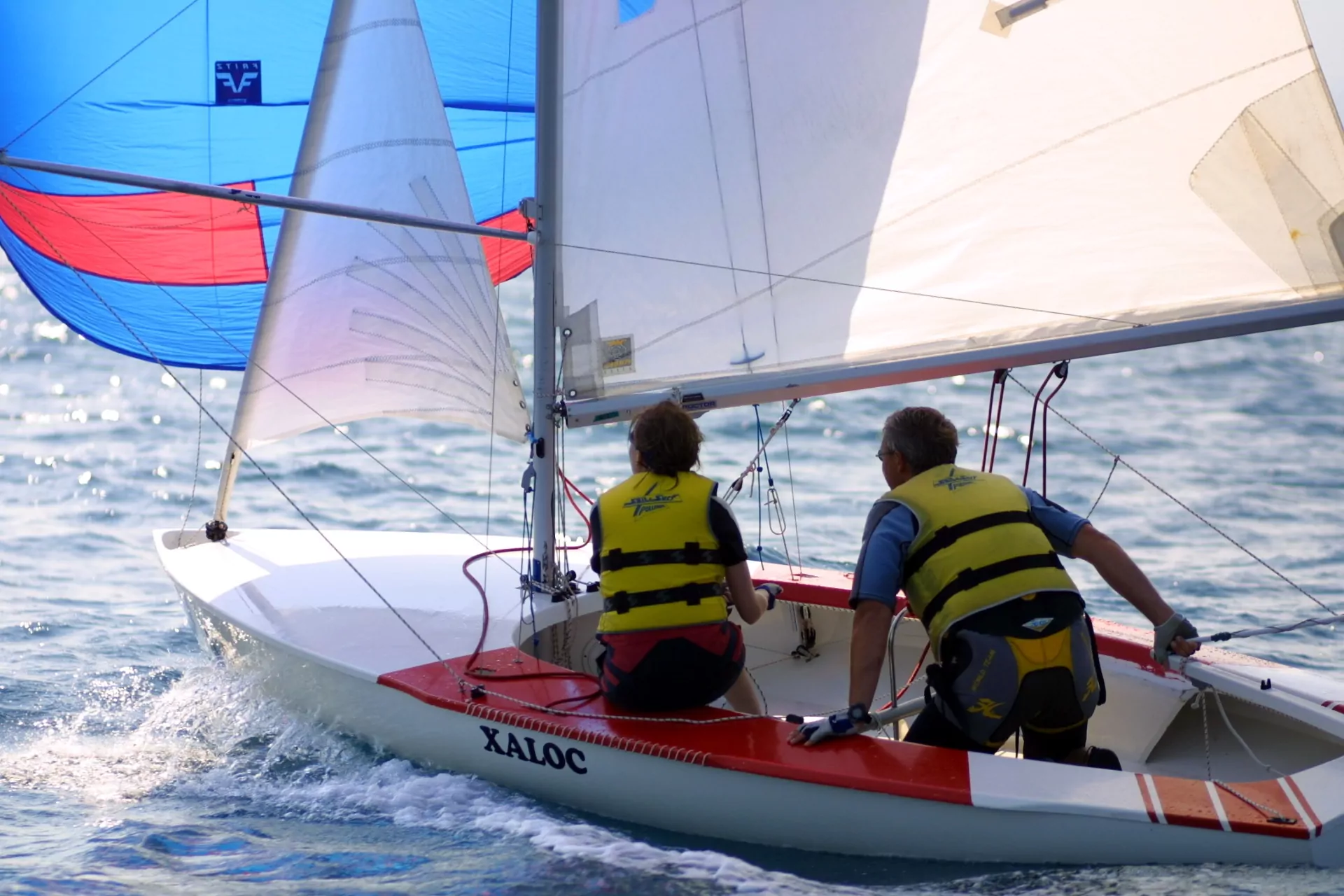 Segelboot mit zwei Personen in gelben Rettungswesten, buntem Spinnaker und weißen Segeln auf sonnigem, glitzerndem Meer
