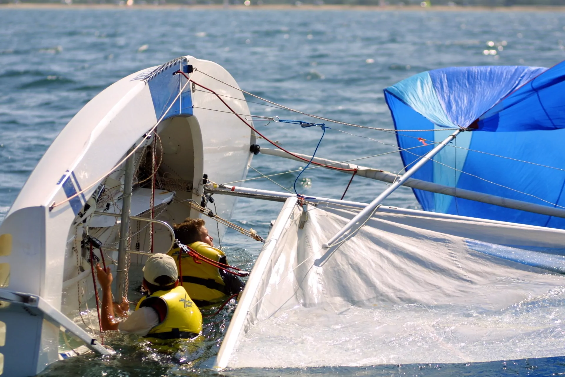 Gekentertes Jollen-Segelboot mit blauem Spinnaker und Leinen, zwei Personen in gelben Schwimmwesten im Wasser