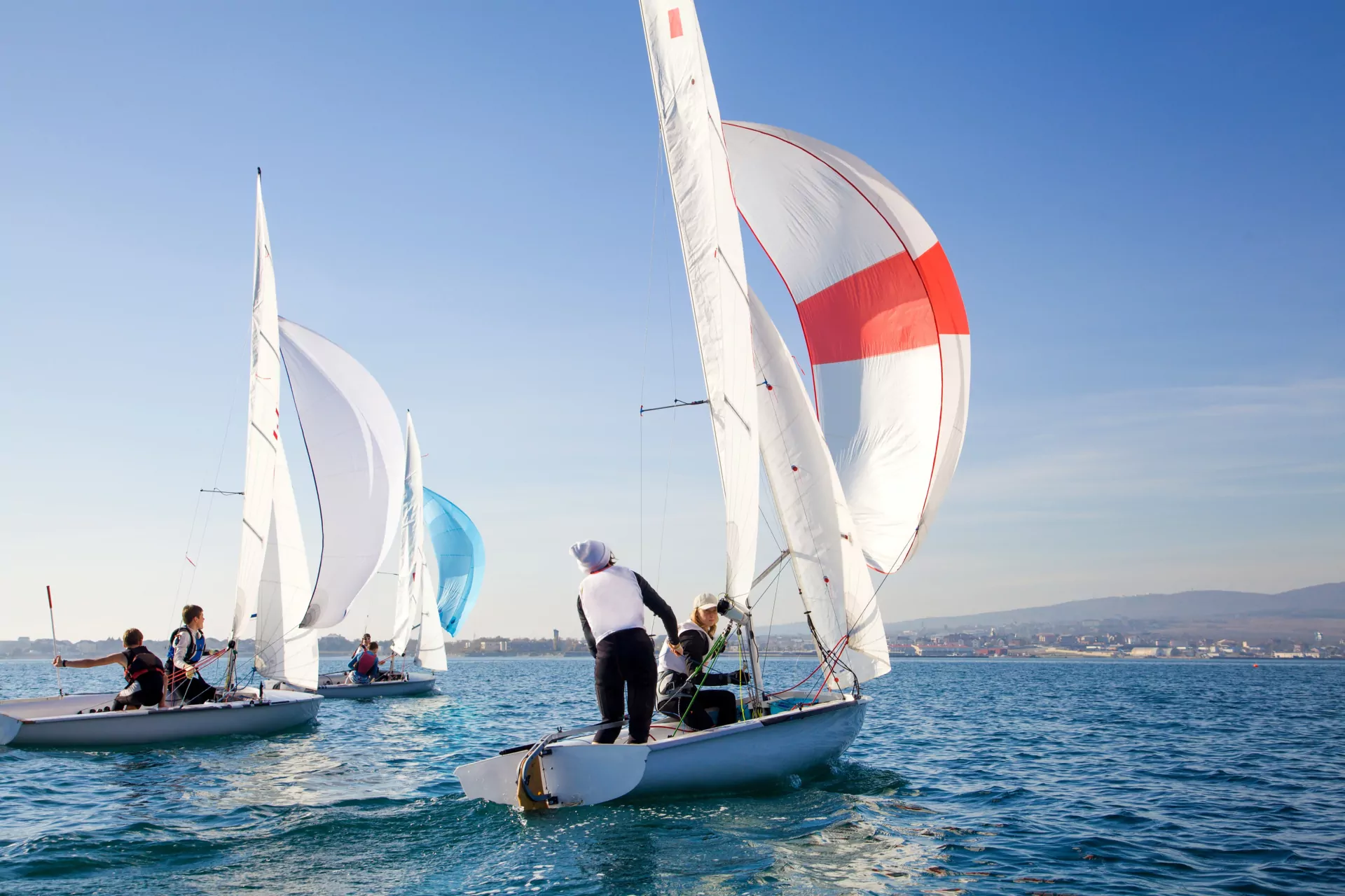 Segelregatta mit mehreren Jollen auf ruhigem Meer, weiße und rote Spinnaker, Crew unter klarem blauem Himmel