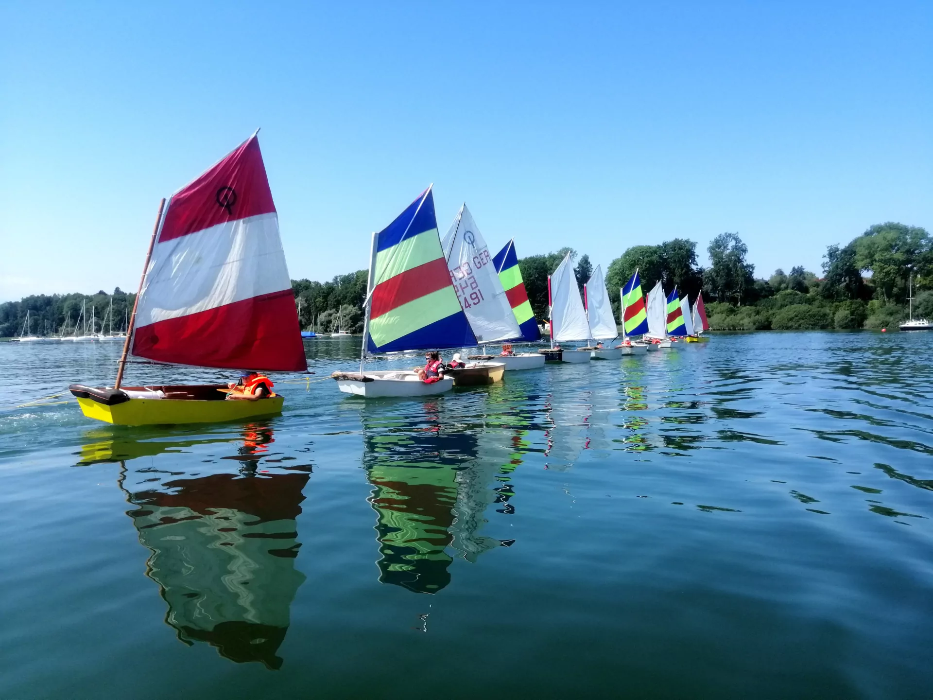 Bunte Segelboote (Optimisten) in Reihe auf ruhigem See bei sonnigem Himmel – Segeltraining/Regatta