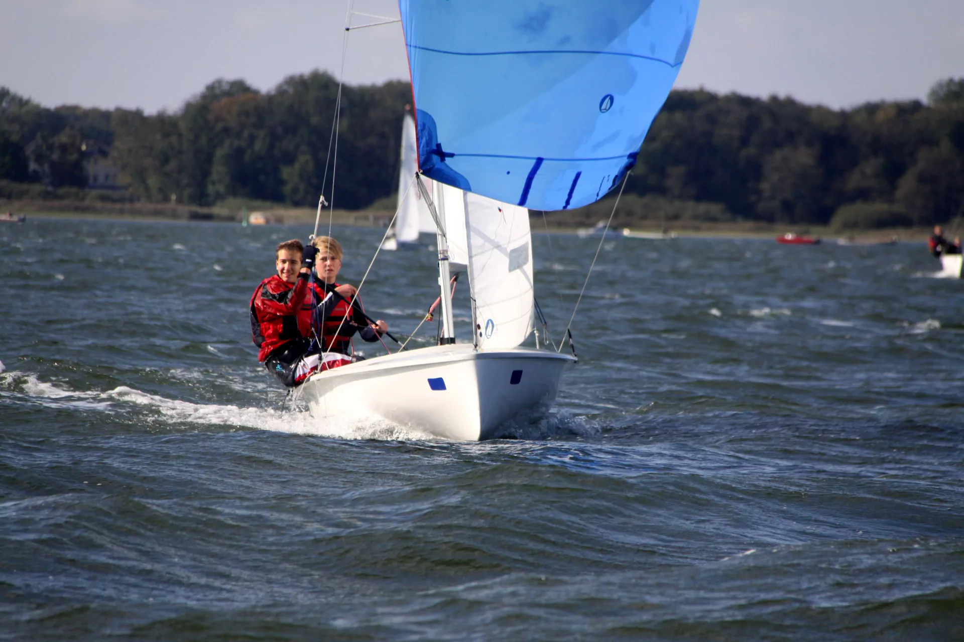 Zwei Jugendliche segeln auf kleinem Jollenboot mit blauem Spinnaker, windige See und Küstenwald im Hintergrund