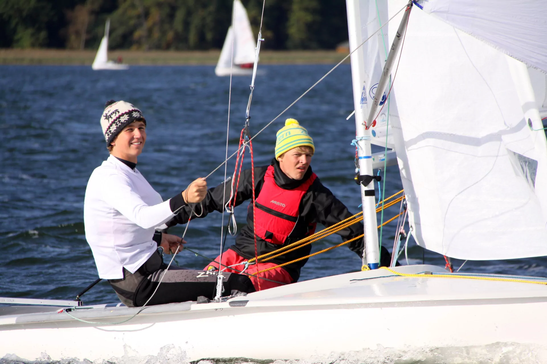 Zwei jugendliche Segler beim Segelsport auf Jolle, rote Rettungsweste, Mützen, weiße Segel auf blauem See