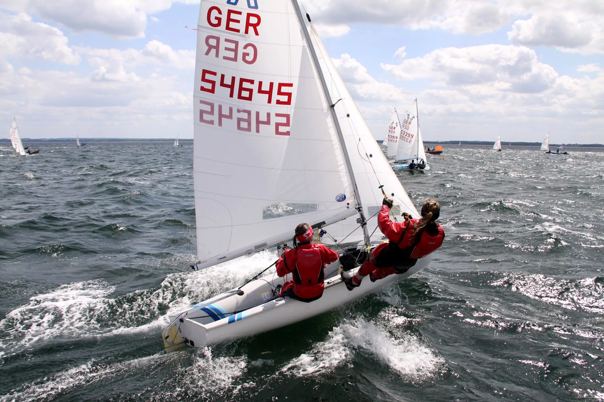 Segelregatta: zwei Segler in roten Anzügen auf Jolle mit GER-Segel, raue Küstenwellen und Sonne