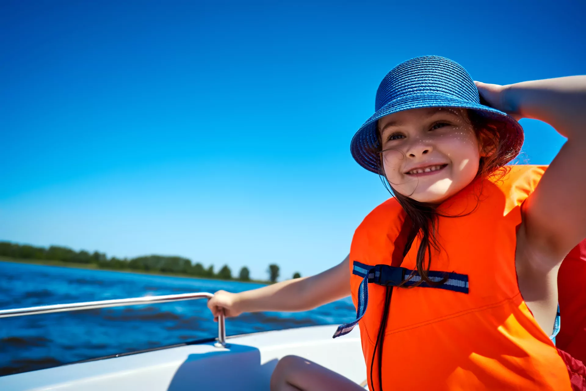 Lächelndes Mädchen mit blauem Sonnenhut und orangefarbener Schwimmweste auf einem Boot am See, sonniger Sommertag