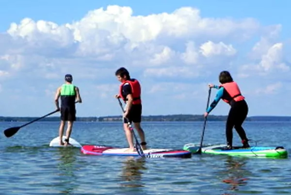 Drei Menschen beim Stand-up-Paddling (SUP) auf einem ruhigen See, mit Paddeln und bunten Rettungswesten, sonniger Himmel