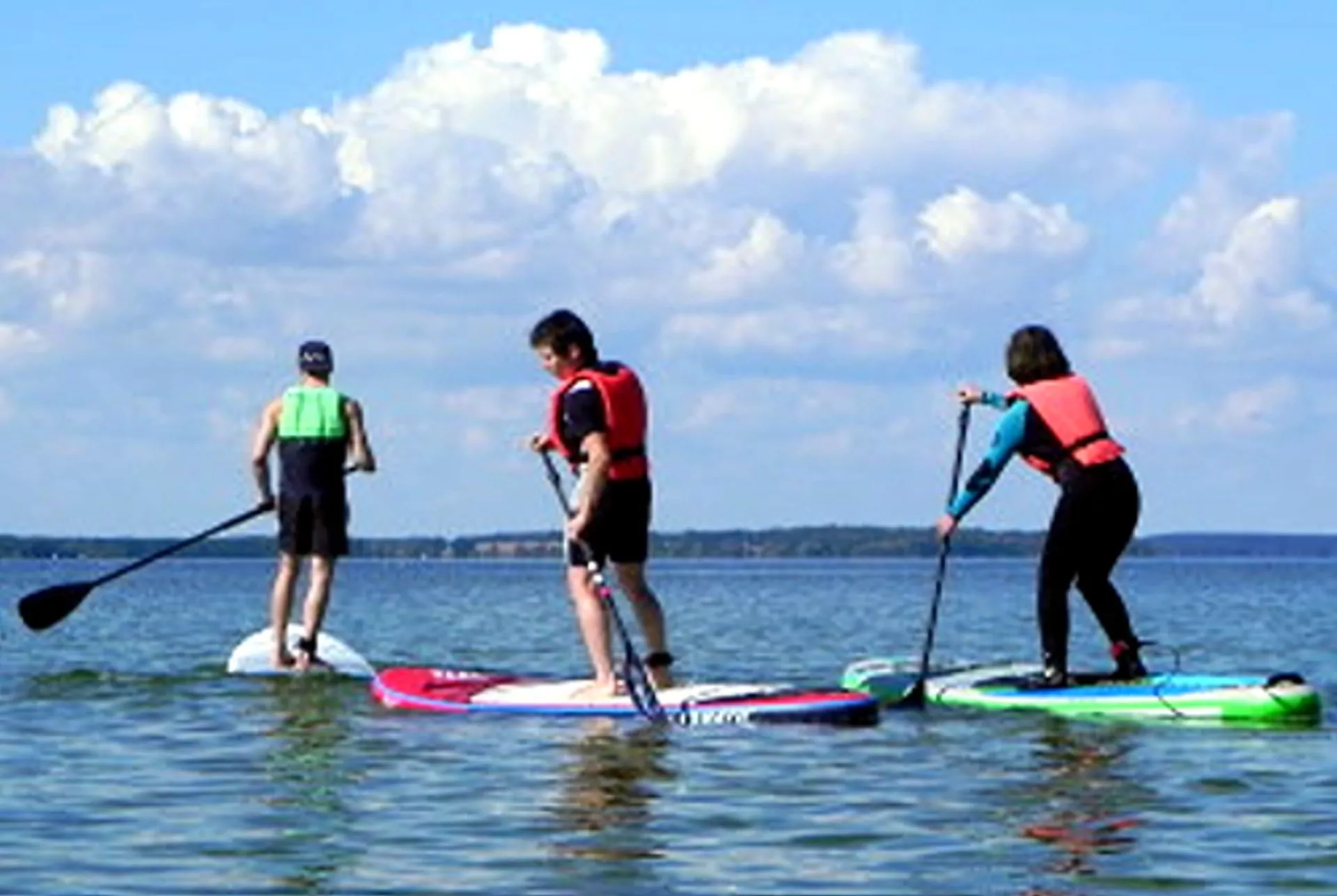 Drei Menschen beim Stand-up-Paddling (SUP) auf einem ruhigen See, mit Paddeln und bunten Rettungswesten, sonniger Himmel