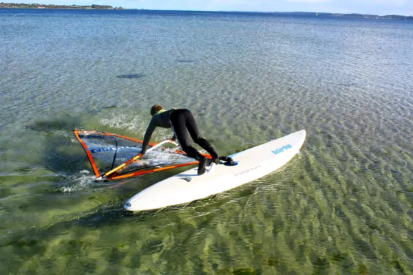 Windsurfer beim Aufstehen auf dem Board in klarem flachem Wasser, orange-blaues Segel, Küstenhorizont im Hintergrund
