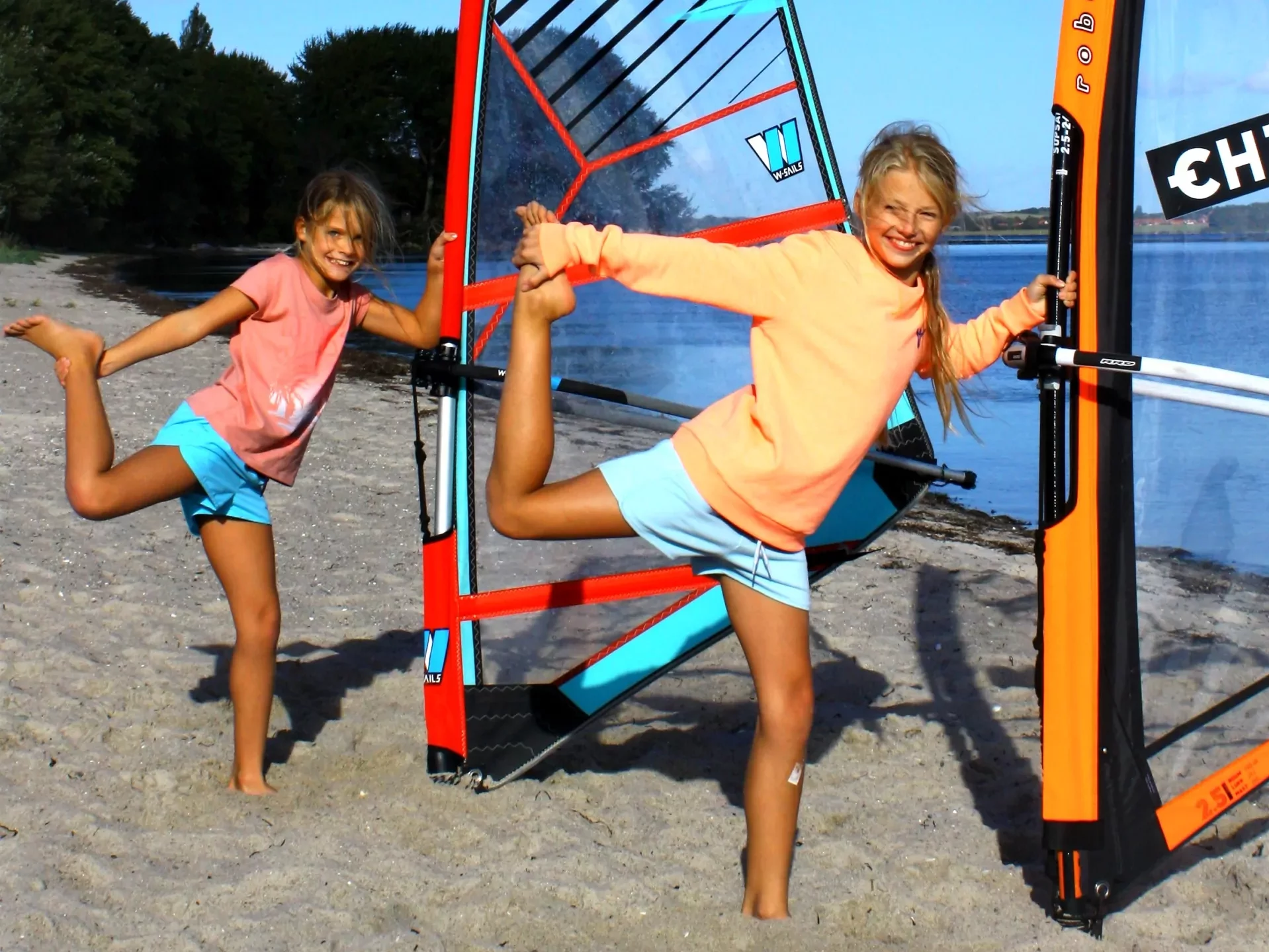 Zwei Mädchen am Strand dehnen sich an bunten Windsurfsegeln, fröhliches Sommersport-Motiv an der Küste
