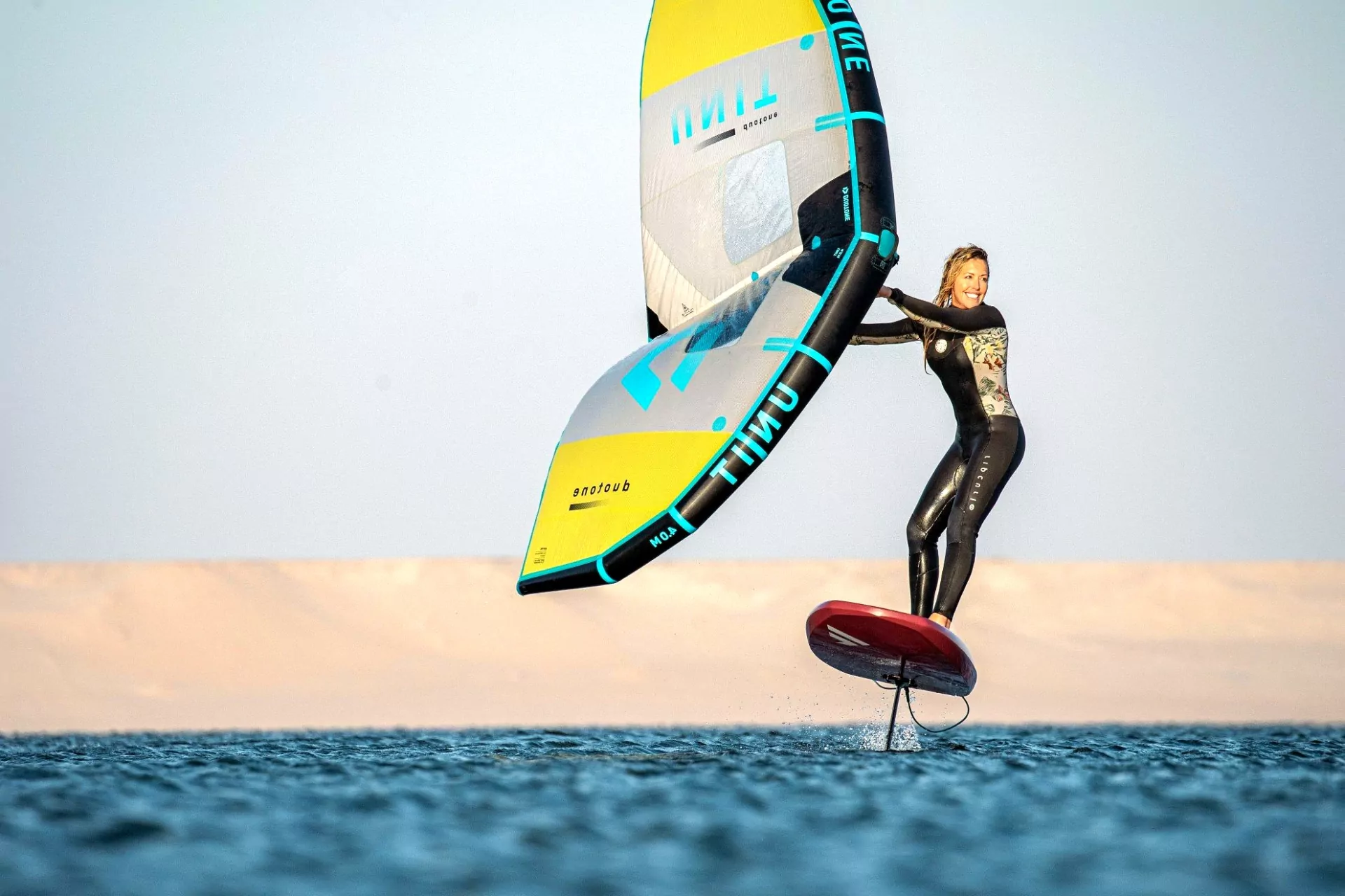 Lächelnde Frau beim Wingfoiling auf Foilboard, gelb-blaues Wing über dem Meer vor hellem Himmel