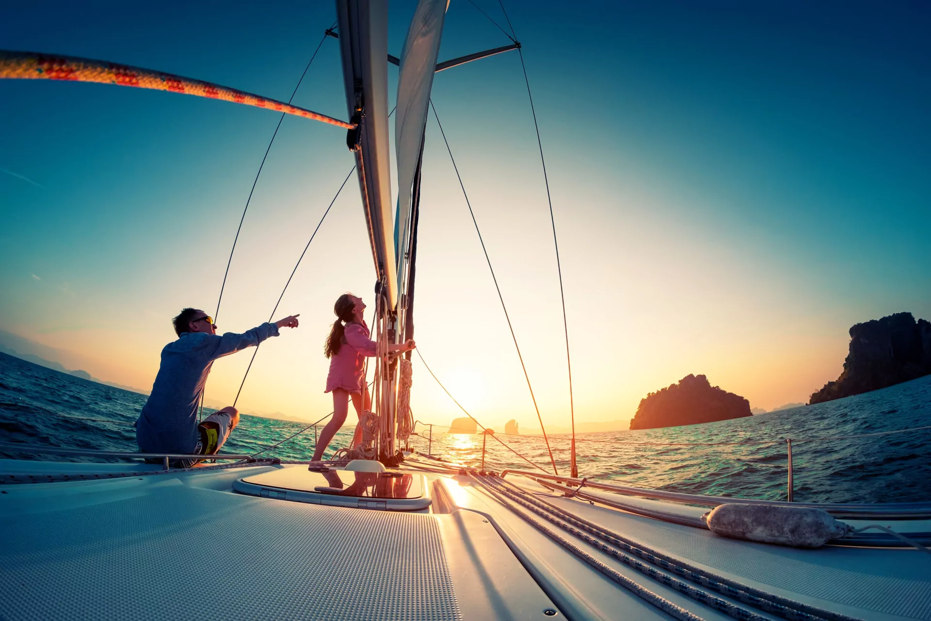 Paar auf Segelboot bei Sonnenuntergang, offenes Meer und Felseninseln im Hintergrund – Segelurlaub