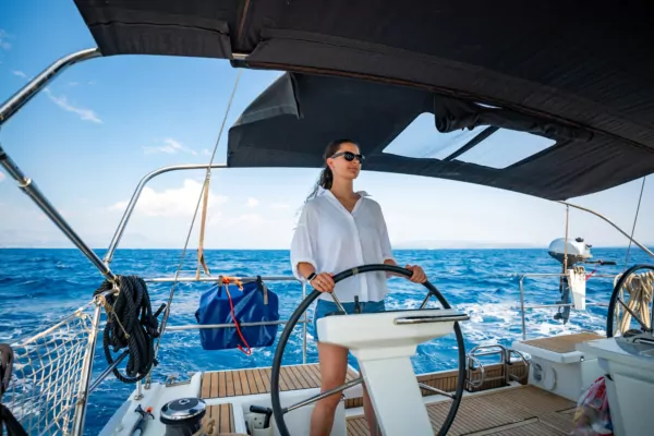 Frau am Steuerrad eines Segelboots, Sonnenbrille, blaues Meer im Hintergrund, Sommersegeln, Freizeit yachting