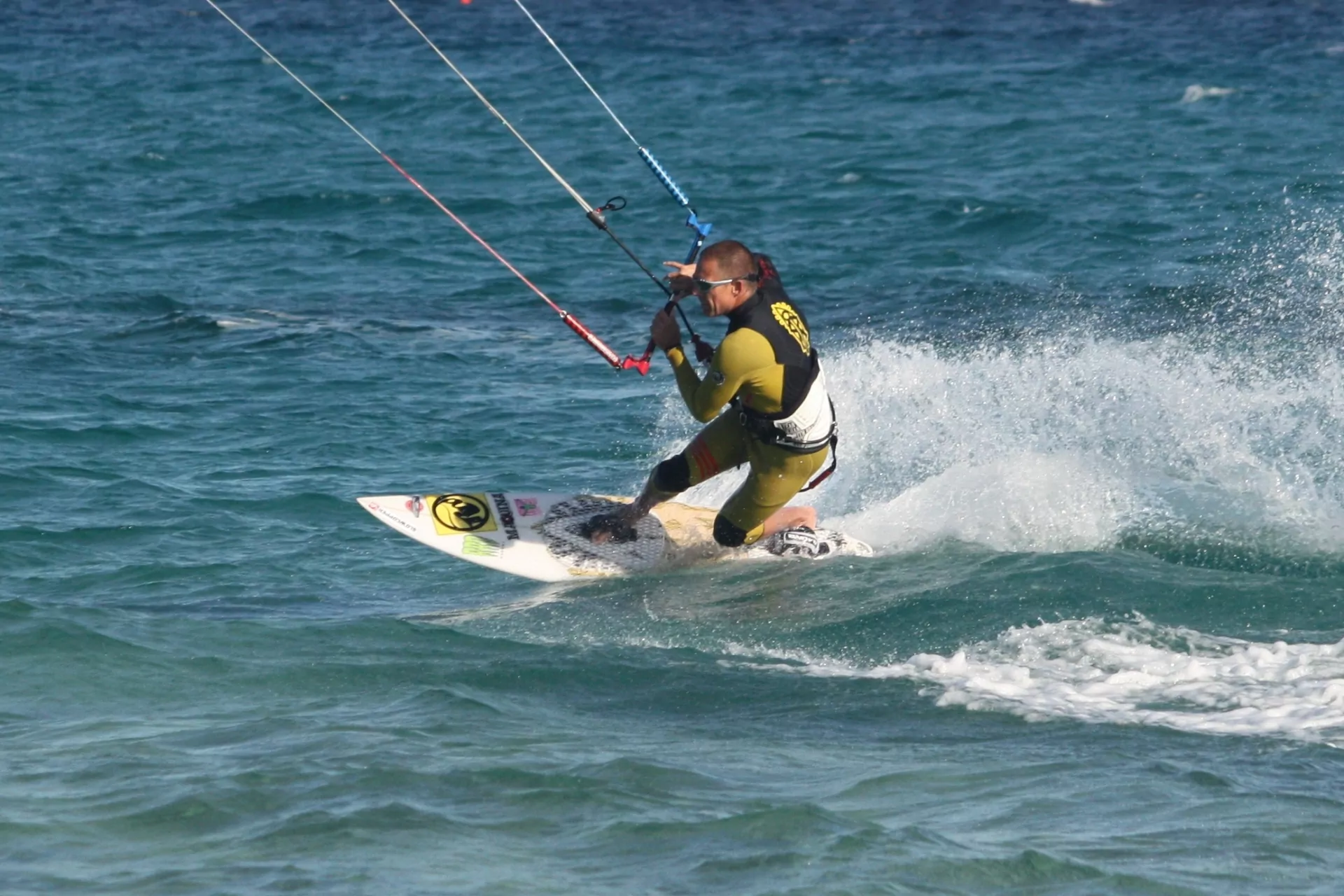 Kitesurfen: Kitesurfer auf weißem Board fährt durchs blaue Meer, erzeugt Spritzwasser – actionreiche Wassersportaufnahme